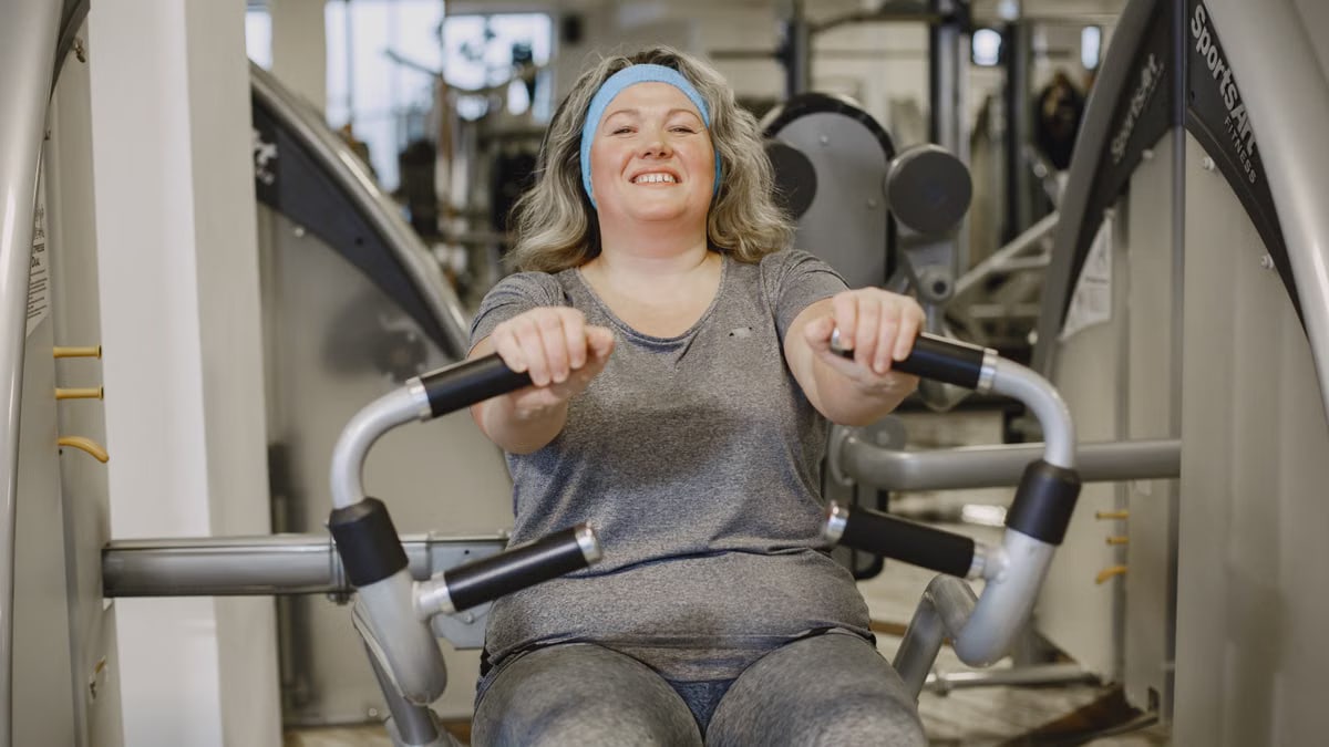 Smiling woman exercises on a gym machine, promoting fitness and well-being.